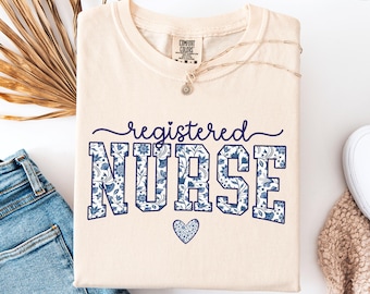 May include: Ivory-colored t-shirt with the words "registered NURSE" in navy blue floral print. A small navy blue heart is below the word "NURSE". The shirt is folded on a white surface next to blue jeans and a beige sweater.