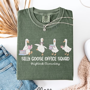 May include: Moss green t-shirt with a graphic of four cartoon geese, each with a different accessory. The text "SILLY GOOSE OFFICE SQUAD" and "Brightoak Elementary" are printed below the geese. A silver necklace is draped on the shirt.