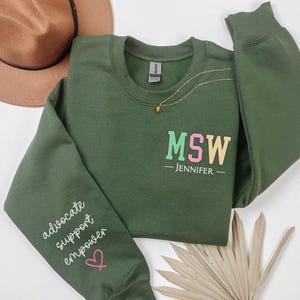 May include: Green sweatshirt with the text "MSW - JENNIFER -" on the front and the text "advocate support empower" on the left sleeve. The sweatshirt has a pink heart on the sleeve.