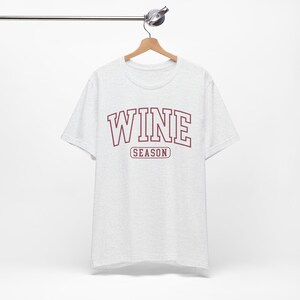 Peut inclure: Un t-shirt gris chin&eacute; avec le mot "WINE" en lettres majuscules bordeaux au-dessus du mot "SEASON" en plus petit. Le t-shirt est suspendu &agrave; un cintre en bois.