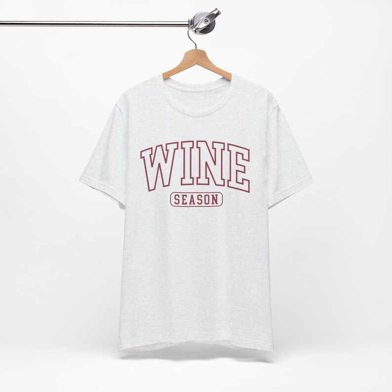 Peut inclure: Un t-shirt gris chin&eacute; avec le mot "WINE" en lettres majuscules bordeaux au-dessus du mot "SEASON" en plus petit. Le t-shirt est suspendu &agrave; un cintre en bois.