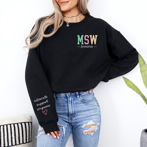 May include: Black sweatshirt with the text "MSW - JENNIFER -" in colorful letters. The left sleeve has the text "advocate support empower" with a pink heart.