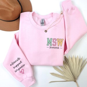 May include: Pink sweatshirt with the text "MSW - JENNIFER -" on the front and the text "advocate support empower" on the left sleeve with a pink heart.