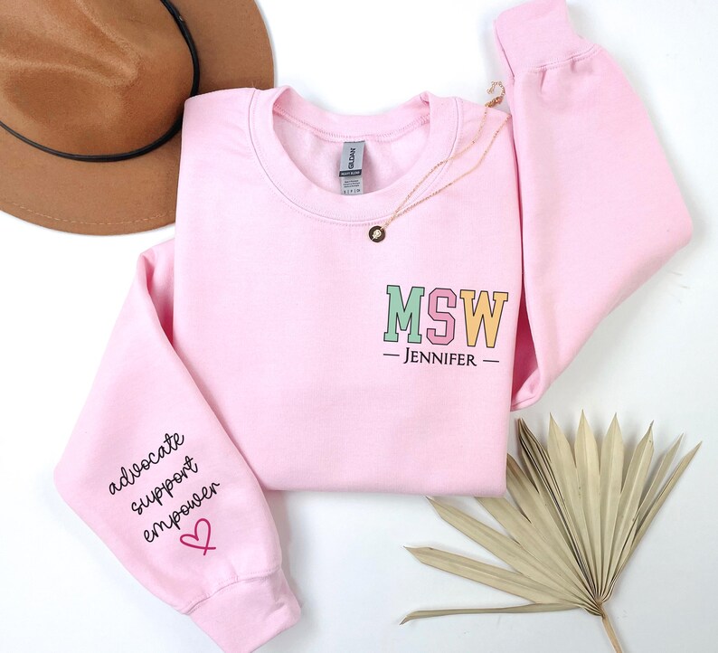 May include: Pink sweatshirt with the text "MSW - JENNIFER -" on the front and the text "advocate support empower" on the left sleeve with a pink heart.