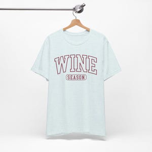 Peut inclure: T-shirt bleu clair avec le mot "WINE" en lettres majuscules bordeaux et le mot "SEASON" en plus petit en dessous. Le t-shirt est suspendu &agrave; un cintre en bois.