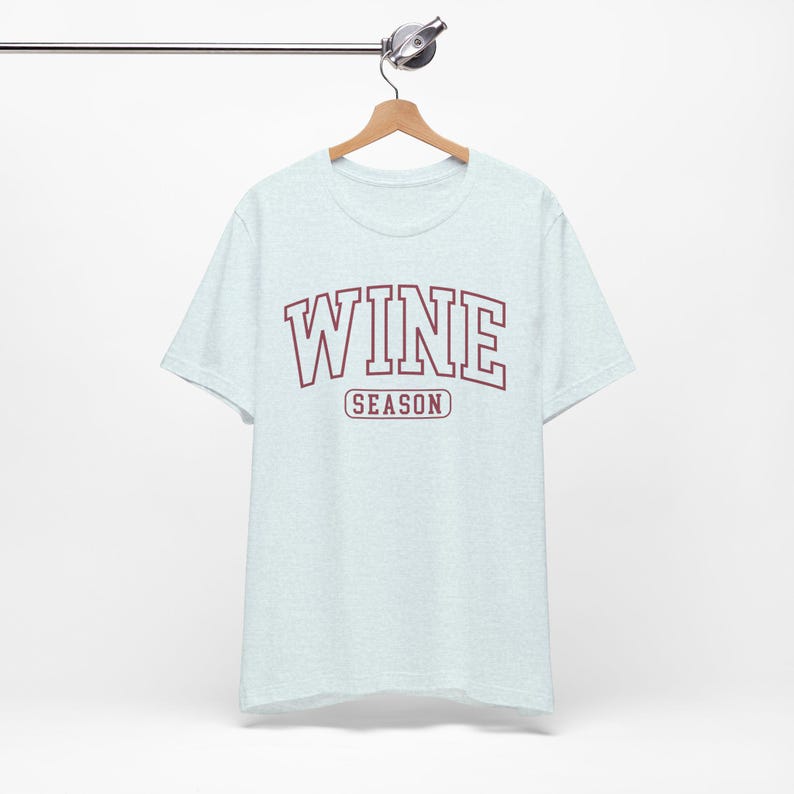 Peut inclure: T-shirt bleu clair avec le mot "WINE" en lettres majuscules bordeaux et le mot "SEASON" en plus petit en dessous. Le t-shirt est suspendu &agrave; un cintre en bois.