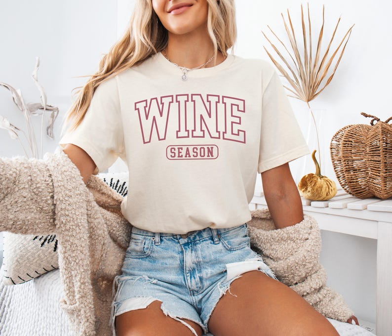 Peut inclure: T-shirt cr&egrave;me avec l'inscription "WINE SEASON" en lettres majuscules bordeaux. Le t-shirt est associ&eacute; &agrave; un short en jean. Un cardigan douillet de couleur claire est drap&eacute; sur les &eacute;paules. Des citrouilles d&eacute;coratives et un panier en osier sont en arri&egrave;re-plan.