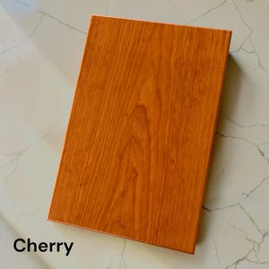 May include: A rectangular cherry wood board with a smooth, warm orange-brown surface featuring a natural vertical grain pattern. The board has straight, clean edges and a flat top surface, displaying the distinct color and texture of cherry wood.