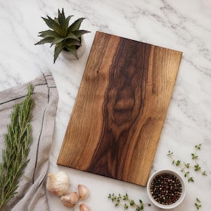 May include: A rectangular wooden cutting board with a prominent dark brown grain pattern. The board is positioned alongside a small potted succulent, a grey linen cloth, fresh rosemary sprigs, garlic cloves, and a small bowl of peppercorns.