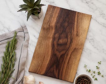 May include: A rectangular wooden cutting board with a prominent dark brown grain pattern. The board is positioned alongside a small potted succulent, a grey linen cloth, fresh rosemary sprigs, garlic cloves, and a small bowl of peppercorns.