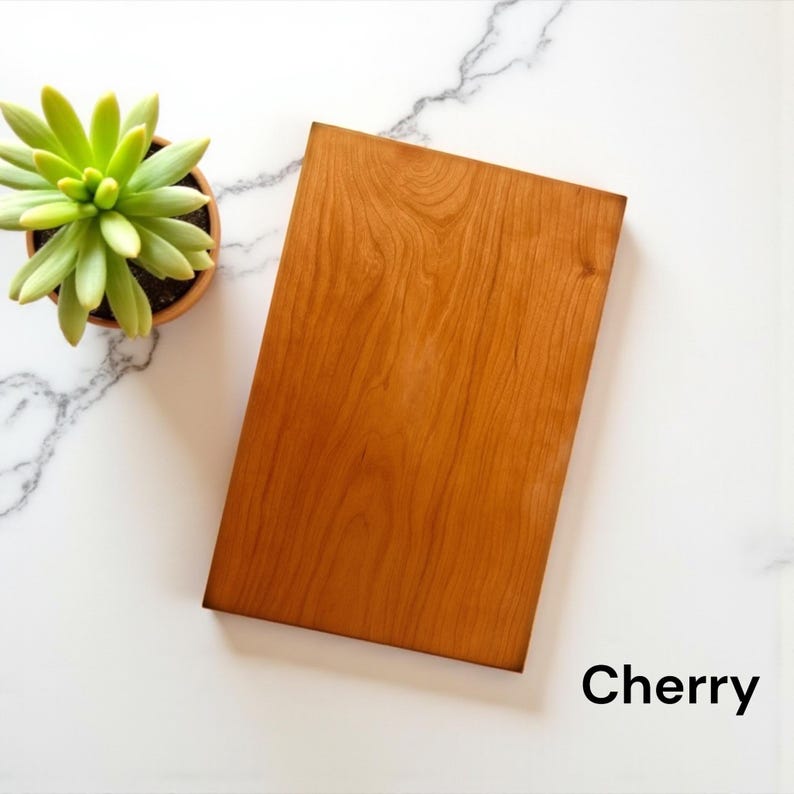May include: A rectangular cherry wood board with a smooth, warm-toned grain pattern. The board is positioned next to a small, light green succulent plant in a terracotta pot. The word Cherry is printed in black text in the bottom right corner.