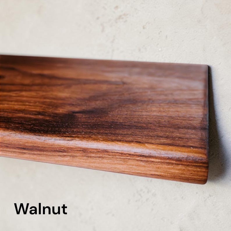 May include: A rectangular shelf made of dark brown walnut wood with a smooth, polished surface and visible natural grain patterns. The wood features deep chocolate and reddish-brown tones throughout the board, with the word Walnut printed below.