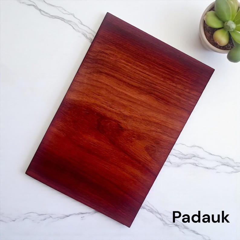 May include: A rectangular Padauk wood board with a deep reddish-brown color and a visible natural grain pattern. The surface is smooth and polished, showcasing the rich, warm tones of the wood. The word Padauk is printed in black text.
