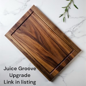 May include: A rectangular wooden cutting board with a dark brown grain pattern and a carved juice groove along the perimeter. The text "Juice Groove Upgrade Link in listing" is printed in black on the lower left side of the image.