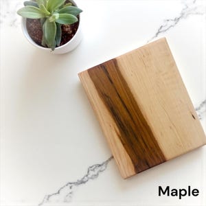 May include: A rectangular wooden cutting board featuring a light cream-colored wood grain with a distinct, dark brown vertical strip running through the center. The word Maple is printed in black text in the bottom right corner of the image.