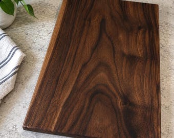 May include: A rectangular wooden cutting board made of dark brown walnut wood with a prominent, swirling natural grain pattern. The board features a smooth, flat surface with straight edges and a deep, rich color palette throughout the wood.