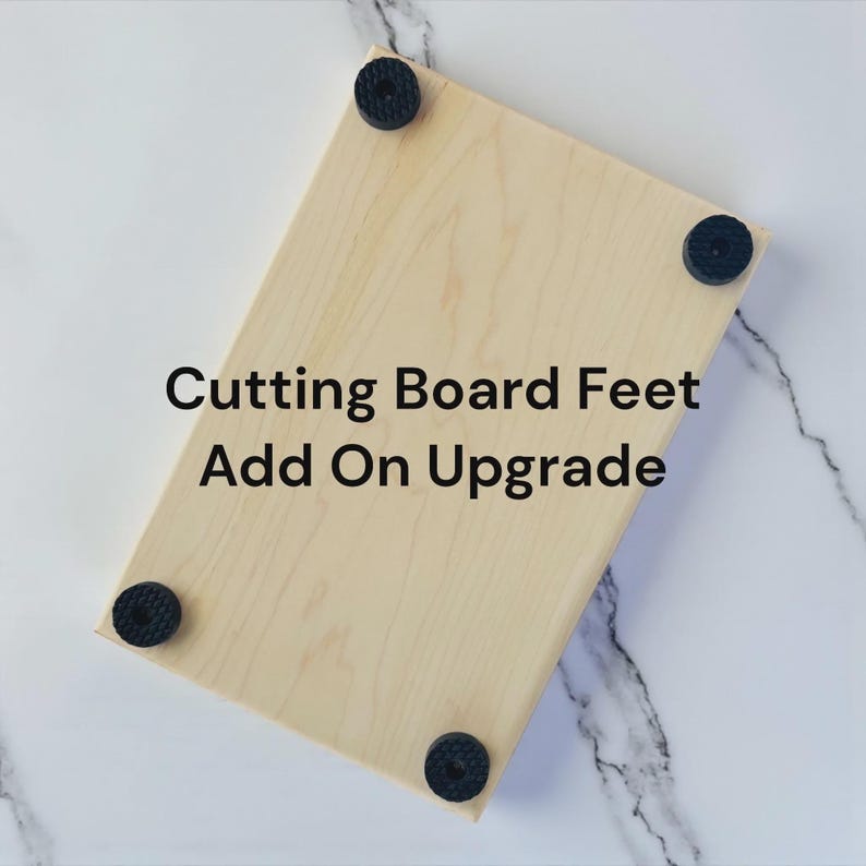 May include: A rectangular light-colored wooden cutting board featuring four circular black rubber feet attached to the corners. The text "Cutting Board Feet Add On Upgrade" is printed in black in the center of the wooden surface.