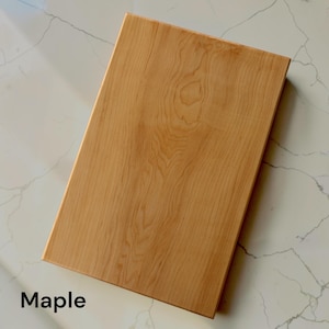 May include: A rectangular wooden cutting board made of light-colored maple wood with a visible natural grain pattern. The board has a smooth, flat surface and straight edges. The word Maple is printed in black text in the bottom left corner.