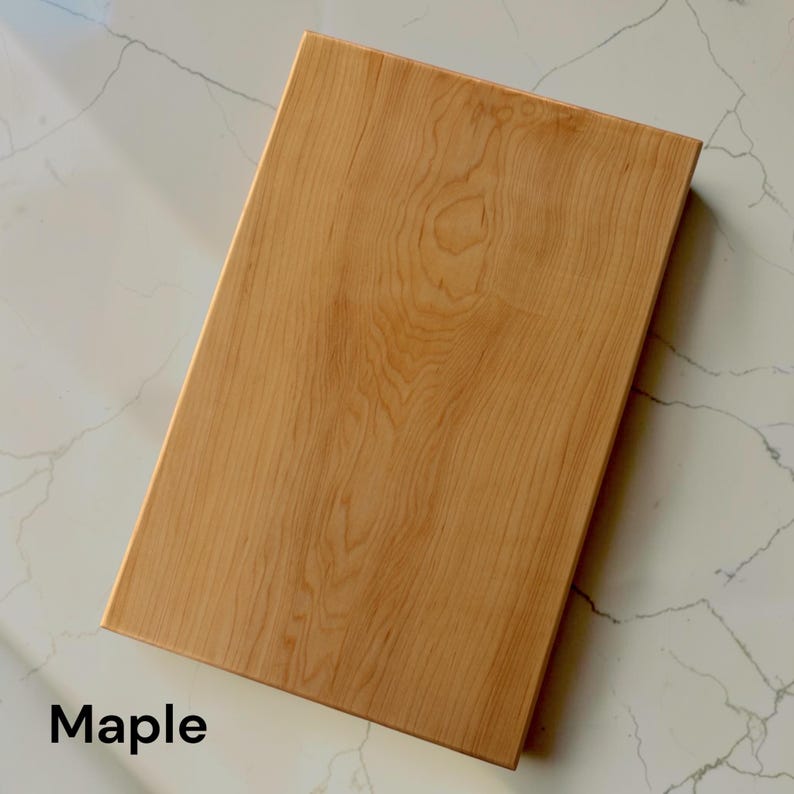 May include: A rectangular wooden cutting board made of light-colored maple wood with a visible natural grain pattern. The board has a smooth, flat surface and straight edges. The word Maple is printed in black text in the bottom left corner.