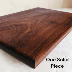 May include: A rectangular dark brown wooden cutting board with a visible natural grain pattern. The surface is smooth and finished. The text "One Solid Piece" is printed in black in the lower right corner of the image, indicating the construction.