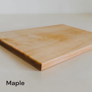 May include: A rectangular light-colored maple wood cutting board with a smooth, sanded surface and visible natural wood grain patterns. The board features straight edges and a flat top surface. The word Maple is printed in black text below.