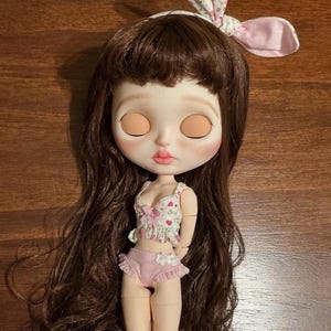 May include: A doll with long wavy brown hair and a white headband featuring a pink floral pattern. The doll has closed eyes, pale skin, and pink lips. It wears a white crop top with a pink heart print and a pink ruffled bottom with white floral accents.