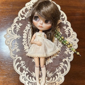 May include: A handmade doll with brown hair, large eyes, and a cream-colored dress. The doll is posed on a cream-colored lace doily. A beaded accessory with green and gold beads is next to the doll.