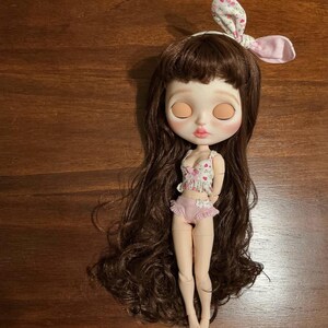 May include: A Blythe doll with long brown hair, wearing a floral print top and ruffled pink bottoms. The doll has a matching floral headband with bunny ears. The doll is standing on a wooden surface.