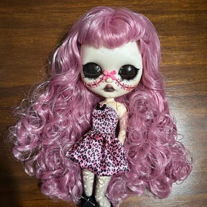 May include: A custom Blythe doll with a unique, artistic design. The doll has large, expressive eyes, stitched facial details, and long, wavy, pink hair. She wears a pink leopard print dress and patterned stockings, with black shoes. The doll is a collectible art piece.