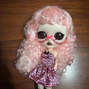 May include: A doll with a pale face, large eyes, and pink curly hair. The doll's face has stitched details and a pink bow. It wears a pink and black leopard print dress with a black bow at the neckline. The doll's legs are visible.