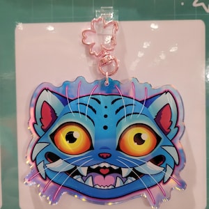 Pode incluir: Um chaveiro azul em forma de gato com um fecho em forma de flor em ouro rosa. O gato tem olhos amarelos grandes, um sorriso largo e orelhas rosa. O chaveiro tem um efeito holográfico iridescente.