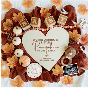 May include: A wooden heart-shaped sign with the text "We are adding a little pumpkin to our patch" and "Arriving November 2024". The sign is surrounded by fall-themed decorations, including small pumpkins, fall leaves, and wooden blocks that spell out "BABY".