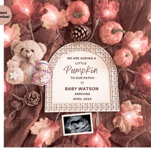 Pode incluir: Uma placa de madeira com um anúncio de bebê com tema de outono. A placa tem a forma de um arco e tem um fundo branco com detalhes marrons. O texto na placa diz "We are adding a little Pumpkin to our patch." A placa também inclui o nome do bebê, "Baby Watson", e a data de chegada prevista, "April 20XX". A placa é cercada por folhas de outono, um urso de pelúcia e uma corda de luzes.