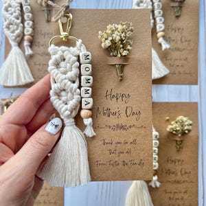 Pode incluir: Um cartão castanho com o texto "Happy Mother's Day" e "Thank you for all that you do! From Foster the Family". Um pequeno buquê de flores secas está preso ao cartão. Um chaveiro de macramé branco com contas de madeira que soletram "MOMMA" está preso ao cartão.