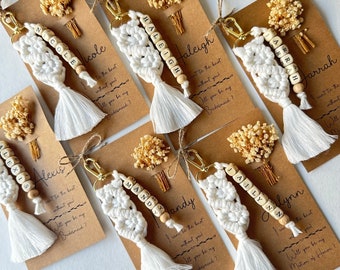 May include: A collection of handmade macrame keychains with wooden bead names, white tassels, and gold-tone hardware. Each keychain is paired with a kraft paper card and dried floral accents. Text on the cards reads, "I can't tie the knot without you! Will you be my Bridesmaid?"