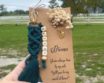 May include: A green and gold tassel keychain with a wooden bead charm that spells "Briana". A card with the text "Briana You've always been by my side. Will you be my maid of honor? 2.2.2024" is attached to the keychain.