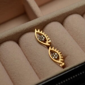 May include: A pair of gold eye-shaped earrings with small clear stones set in the center of each eye. The earrings are in a velvet-lined jewelry box.