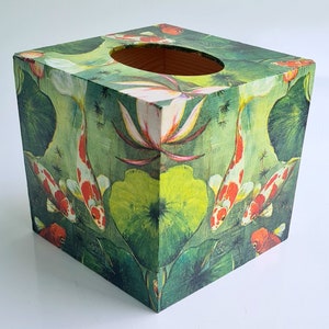 Pode incluir: Tampa quadrada para caixa de lenços com um design floral verde, vermelho e branco com peixes koi nadando em um lago. A caixa é de madeira e tem uma fenda para lenços.