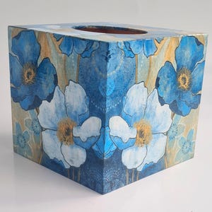 Könnte beinhalten: Quadratischer Taschentuchbox-Bezug mit Blumenmuster. Das Muster zeigt blaue und weiße Blumen auf beigefarbenem Hintergrund.