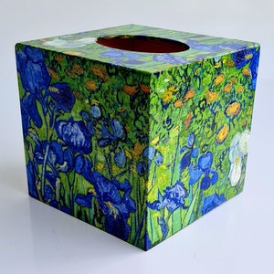 May include: A square tissue box cover with a colorful floral design. The design features blue irises and yellow flowers on a green background.