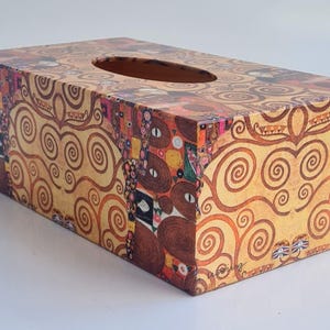 May include: Rectangular tissue box with a decorative design featuring swirling patterns in gold, brown, and red. The box has an oval opening for tissue dispensing. The artwork is reminiscent of the style of Gustav Klimt.
