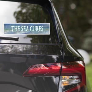 Peut inclure: Un autocollant rectangulaire avec une bordure blanche présentant un fond de vagues océaniques dans des tons de bleu, vert et blanc. Le texte THE SEA CURES est imprimé dans une police serif blanche, stylisée et en gras au centre de l'autocollant.