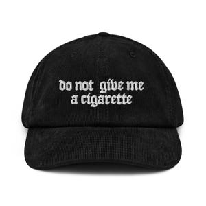 Könnte beinhalten: Schwarze Cord-Baseballkappe mit dem weißen, gestickten Text "do not give me a cigarette". Die Kappe hat einen gebogenen Schirm und eine strukturierte Krone. Ein klassisches Accessoire.