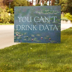 May include: A rectangular yard sign with the text "YOU CAN'T DRINK DATA" in white letters. The sign features a painting of water lilies in a pond. The sign is placed in a grassy yard.