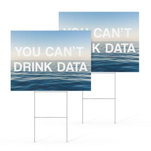 May include: Two yard signs with the text "YOU CAN'T DRINK DATA" and "YOU CAN'T K DATA" in white block letters. The background of the signs is a seascape with blue water and a light blue sky. The signs are mounted on metal stakes.