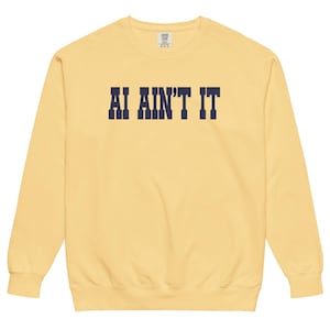 Puede incluir: Una sudadera de cuello redondo en color amarillo pálido con el texto AI AIN'T IT impreso en letras de bloque azul marino oscuro en el pecho. La prenda tiene mangas largas, cuello acanalado y un corte estándar con etiqueta de marca visible.