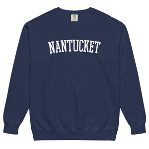 May include: Navy blue sweatshirt with the word "NANTUCKET" in large, white, block letters across the chest. The sweatshirt has a crew neck and long sleeves, and is made of a soft material.