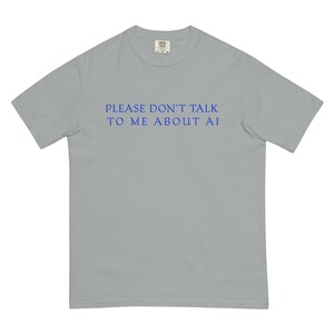 May include: A light gray t-shirt with the text "PLEASE DON'T TALK TO ME ABOUT AI" in blue. The shirt has short sleeves and a crew neck. The text is centered on the front of the shirt.