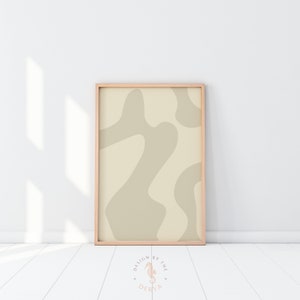 May include: A framed abstract art print featuring a beige and light tan organic design. The artwork is in a light wood frame and leans against a white wall and floor. The print has a minimalist aesthetic.
