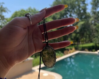 May include: A green and white stone pendant on a black cord necklace. The pendant is oval shaped and has a natural, organic look. The necklace is hanging from a person's hand.